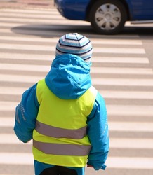 Little boy is crossing zebra crossing where any reckless driver of car pulled into. Child is wearing yellow reflective vest and jacket with reflective strips because of safety.