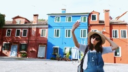 VENICE, BURANO, ITALY - JULY 7, 2018: Burano Island with multicolor houses near Venice. tourist, young woman, girl in sun glasses, hat, backpack, makes selfie with smartphone against the background of