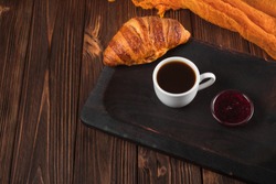 Freshly baked croissant with cup of coffee on brown wooden background. Fresh pastries for breakfast in modern dark mood style.