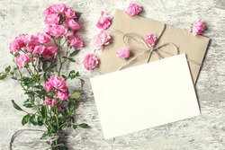 blank white greeting card with pink rose flowers bouquet and envelope with flower buds on white wooden background. vintage toning.top view. mock up