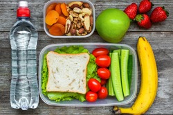 Healthy lunch boxes with sandwich and fresh vegetables, bottle of water, nuts and fruits on rustic wooden background. top view
