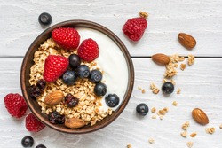 bowl of oat granola with yogurt, fresh raspberries, blueberries and nuts on white wooden board for healthy breakfast. top view