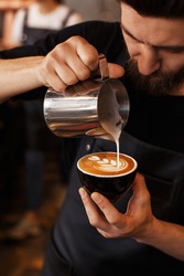 Young smiling male barista making cappuccino. Pouring milk for prepare cup of coffee. Latte art. Morning cup of coffee in café. Brewing coffee. Coffee shop concept.