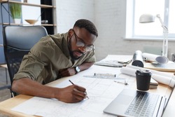 Afro-American architect working in office with blueprints.Engineer inspect architectural plan, sketching a construction project. Portrait of black handsome man sitting at workplace. Business concept.