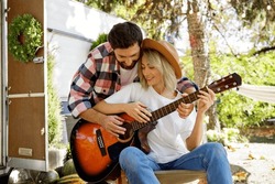 Funny and cheerful young couple. The guy plays the guitar, sing and dance together against the backdrop of a travel trailer. Rest outside the city. Happy people enjoy. Weekends in the summer time.