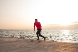 skater in red shirt and blue jeans riding on concrete pier on longboard during sunrise, sea or ocean background 