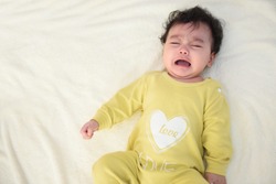 Top view of a cute little Asian baby girl wearing yellow dress, lying on the bed, sad and crying