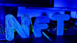 NFT ( Non-Fungible Token ) text of letters illuminated with blue neon light. Workplace of a graphic designer creating NFT digital art