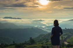Woman standing with her hand in the pocket on the mountain top looking at panoramic view of mountain range in the mist under the sunrise