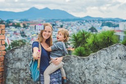 Young woman and her son taking smart phone self portrait pictures with selfie stickon the background of the city of Dalat, Vietnam.