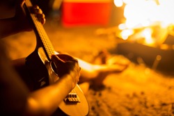 A person relaxing while sitting next to a campfire on a beach, playing a guitar