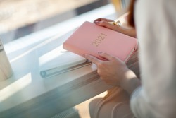 Female hands holding pink or coral coloured leather diary 2021 and pen while sitting near a window Concept of planning personal future goals and ideas for new year 2021. Copy space.