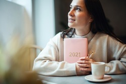 Inspired young woman with a smile looking through window and holding coral colored diary 2021. Hope and inspiration concept. Lady is smiling and dreaming about future new year. Happiness and success.