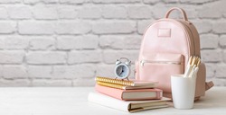 Female school backpack with stationery supplies on desk at white loft brick background. Elegant bag for carrying schooler accessories with notebook, pencil and sketchbook. Back to school concept