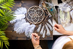Top view female craftsman hands creating traditional dreamcatcher decor choosing material at workshop. Woman artist making native tribal spiritual amulet use natural feathers, beads and thread
