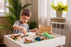 Cute baby boy playing sensory box dinosaur world kinetic sand table with carnivorous and herbivorous dinosaurs. Male kid enjoying early development game fine motor skills with Montessori material