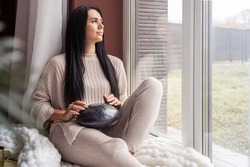 Smiling young woman stroking playing glucophone metal tongue drum sitting on windowsill relaxing at home. Happy female enjoying melody audio tool for meditation, relaxation with deep magical sound