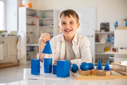 Portrait happy male kid posing with wood Montessori blue geometrical solid volumes figure at classroom. Alternative educational wooden material geometry mathematical volumetric size measurement shape