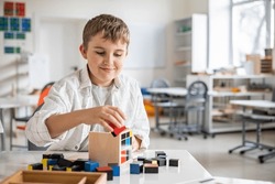Happy male kid assembling Montessori material trinomial cube at school classroom desk. Smiling child boy learning mathematic algebra geometry alternative educational method construction wooden block