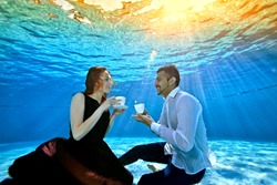 An unusual loving couple, a guy and a girl, look at each other, sitting underwater at the bottom of the pool with white cups in their hands. Surrealism. The concept of tranquility and serenity