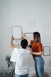 Excited african young husband and caucasian wife hang frame picture on wall happy settling together moving to new house, overjoyed biracial couple decorating own home relocating, ownership concept