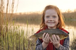 happy redhead girl with smile holding catch freshwater crucian carp fish in hands near lake on weekend