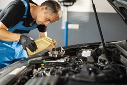 The mechanic is pouring oil into the engine. Pouring fresh oil to car engine, oil change to auto. Caucasian man in blue overalls pouring oil from plastic container.