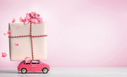 Pink toy retro car with gift box on a roof and flowers on pink background. Copy space.
