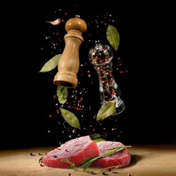 Raw beef steaks and spice mills with peppercorns and herbs flying over table on black background