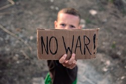 A boy with a grimy face and sad eyes holds a cardboard poster with the inscription NO WAR in English in his hands. A small boy of European appearance opposes military action.