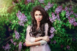 Spring fashion girl outdoors portrait in blooming trees.Beauty romantic woman in flowers of lilac.Sensual lady with long dark shiny hair. Beautiful girl in lacy dress looking at camera. Warm photo.