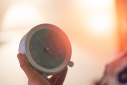Asia woman on bed head on pillow hand holding white teal round alarm clock at 5.00 a.m. in morning as bio clock usually wake up on time with light effect in bedroom. Biological Clock Day in April 28th