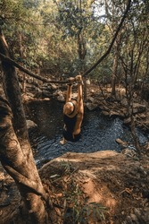 a young woman in black shorts and a straw hat hangs from a branch and swings to jump off the top into a natural pool in a stream in the middle of nature