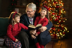 Grandfather wearing glasses, reading a book to small granddaughters twins in a room decorated for Christmas on the background of a Christmas tree. Christmas holiday concept. contrast photography