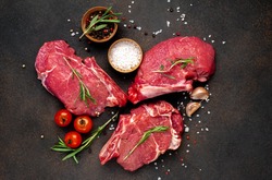 
raw three beef steaks on a cutting board with spices on a stone background