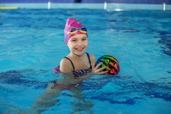 Happy child schoolgirl is studying at swimming lessons in pool. Swimming cap and goggles. Lots of colorful balls. Concept of healthy lifestyle and sports.