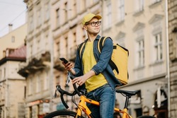A happy delivery man is sitting on a bicycle and texting on his smartphone. He is carrying a yellow backpack for delivery on his shoulders and cap. He is looking around.