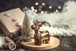 Christmas festive background with toy deer with a gift box, blurred background with golden lights, festive background on wooden deck table and cozy sweater on background