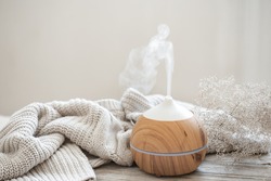 Aroma composition with a modern aroma oil diffuser on a wooden surface with a knitted element and a sprig of dried flowers.