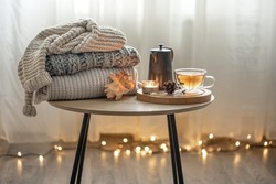 Home autumn composition with tea and knitted sweaters in the interior of the room, on a blurred background with a garland.
