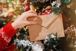 Children's hand holds an envelope with a letter to Santa on Christmas Eve against the background of a decorated Christmas tree with bright lights. Selective focus.