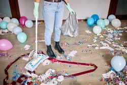 Woman with pushbroom cleaning mess of floor in room after party confetti, morning after party celebration, housework, cleaning service