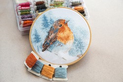 view of the cross stitch process in the hoop multicolored bird, white canvas, floss threads on bobbins, needle, needlework and cross stitch concept 