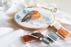 cross stitch process in the hoop multicolored bird, white canvas, floss threads on bobbins, needle, needlework and cross stitch concept 