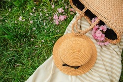picnic blanket with straw hat and bag on green grass covered with pink sakura flowers copy space