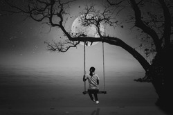 A black and white photo of a woman sitting on a swing and watching the full moon by the sea alone.