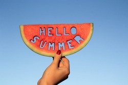 Watermelon slice  with text Hello Summer,  woman hands holding it against blue sky. Summertime concept.