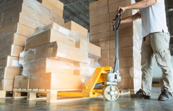 Young man with hand pallet truck or pallet jack and stack of cardboard boxes on pallet in distribution warehouse.