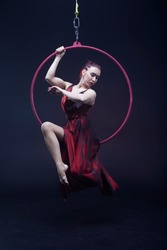 Circus performer woman in red dress doing tricks on red Lyra isolated on black background.