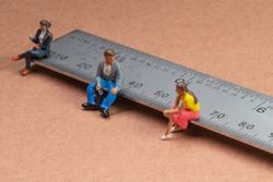 Miniature figures sit on a metal rule or ruler demonstrating the concept of social distancing to avoid transmission of the corona virus or other contagious illness